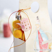 Geschenktüte mit gelben Pompons, einem rosa Teekannenanhänger und einem kleinen Tassenanhänger aus dem Alexandra Renke Motivstanze Kleines Teeset, gebunden mit orangefarbener Schnur.