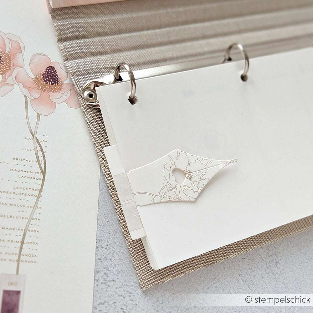 Offene Mappe mit Blumenpapier und einer dekorativen Alexandra Renke Motivstanze Lesezeichen Feder, daneben ein bemaltes Blumenblatt - ideal zum Organisieren von Bastelarbeiten mit dem typischen Alexandra Renke Charme.