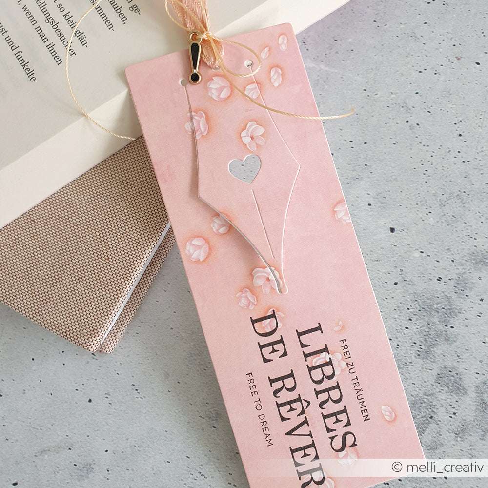 Das rosafarbene Lesezeichen von Alexandra Renke zeigt ein florales Design, eine goldene Schnur und den Text "Libres de rêver". Das Lesezeichen wurde mit tiefem Motivstanzen gefertigt und liegt schön auf einem aufgeschlagenen Buch.
