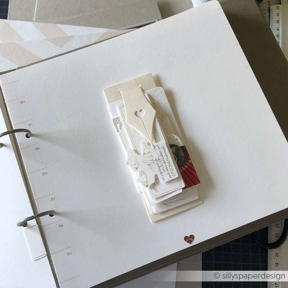 Eine Scrapbook-Seite mit geschichteten Papierverzierungen und Anhängern in neutralen Tönen, die mit Alexandra Renke Deep Edge Motivstanzen und PAPPcut®ies gestaltet wurde, liegt offen auf einem Schreibtisch.