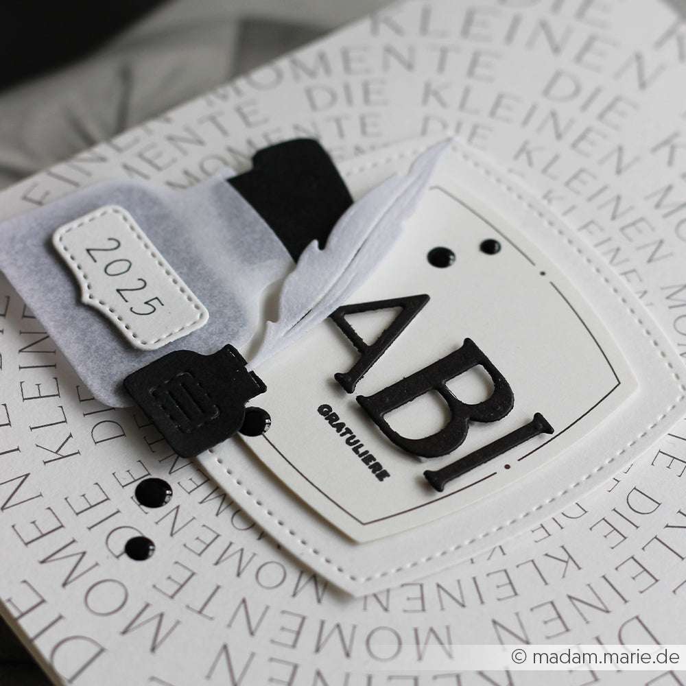 Eine Abschlusskarte von Alexandra Renke mit dem Schriftzug "ABI", einer Feder, einem Tintenfass und einem Anhänger mit der Nummer 2025 auf einem gemusterten Hintergrund, der mit Motivstanze oder Deep Edge Stanzen für komplizierte Akzente gestaltet wurde.