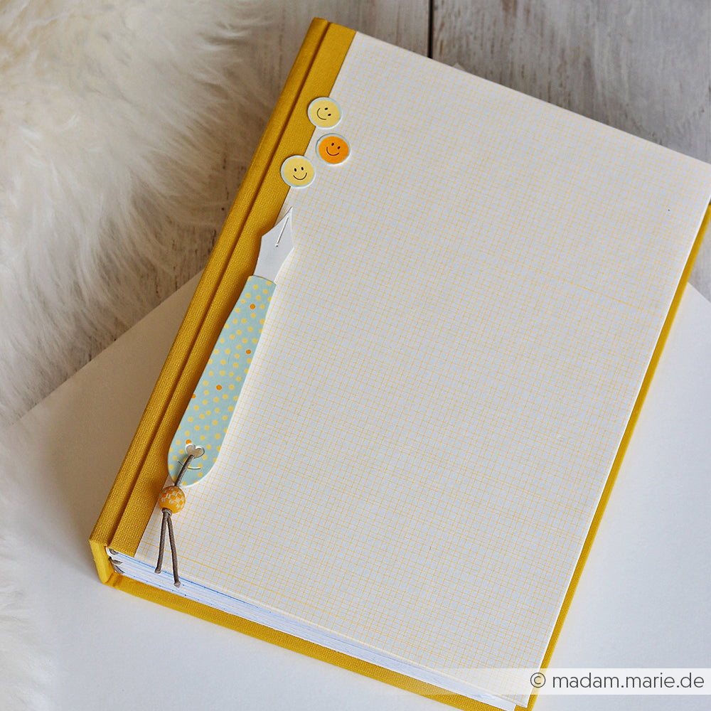 Ein gelbes Notizbuch mit einem Stift, einer Büroklammer und drei Smiley-Buttons auf dem Einband, das auf einer Holzunterlage liegt – eine verspielte Note, die durch PAPPcut®ies und Deep Edge Motivstanzen-Motive noch unterstrichen wird.