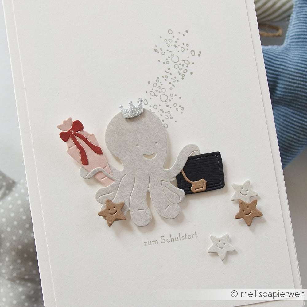 Die niedliche Karte von Alexandra Renke zeigt einen lächelnden Oktopus mit Aktentasche und Schultüte, umgeben von Seesternen. Hergestellt mit Deep Edge Motivstanzen aus dem PAPPcuties Kleines Einschulungs-Set. Inklusive charmantem deutschen Text.
