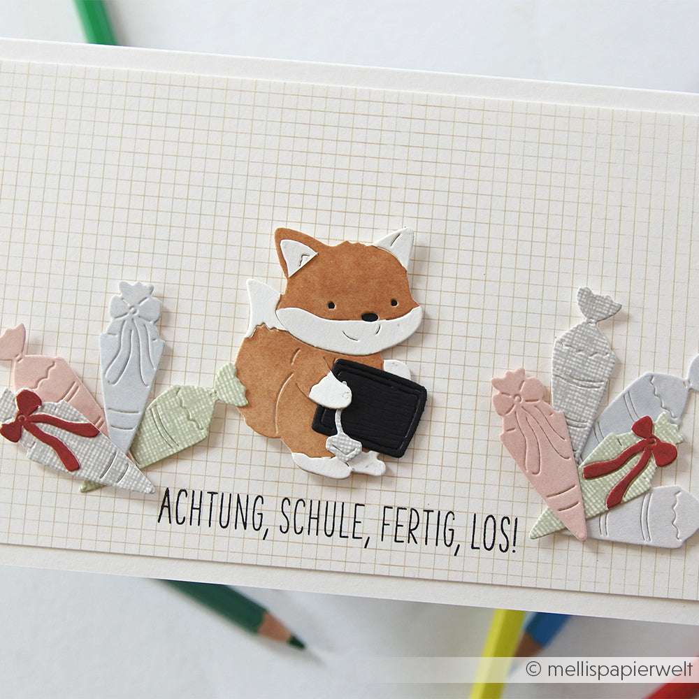 Ein Papierfuchs aus der Serie PAPPcuties von Alexandra Renke hält eine Kreidetafel zwischen Schultüten aus Papier, die mit Deep Edge Motivstanzen auf einem gerasterten Hintergrund mit deutschem Text darunter gestaltet wurden.