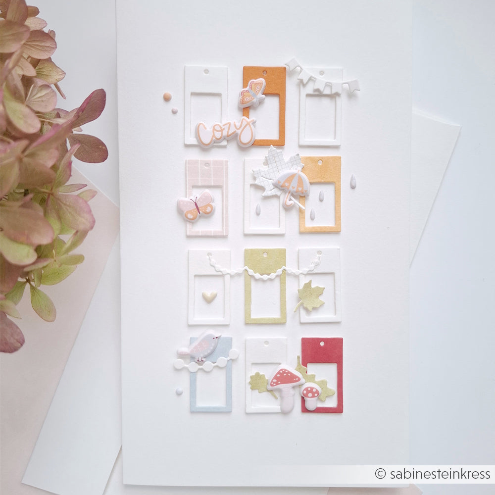 Eine weiße Karte mit zwölf pastellfarbenen Fensterrahmen, Herbstdekorationen und Blumenakzenten, die sorgfältig angeordnet sind – mit den PAPPcut®ies Deep Edge Motivstanzen bis ins kleinste Detail veredelt, was ihr ein klares und elegantes Aussehen verleiht.
