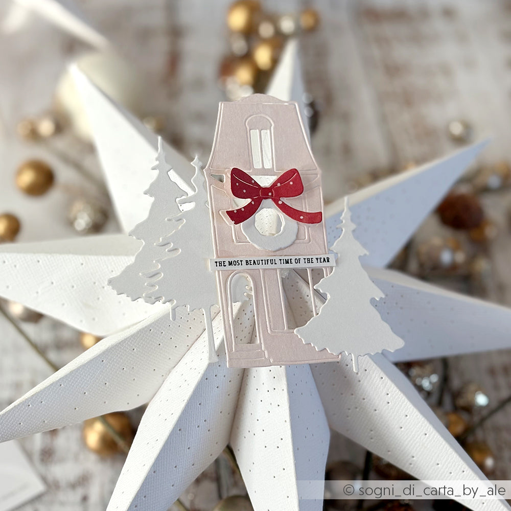 Haus und Bäume aus Papier von Alexandra Renke Designpapiere mit der Motivstanze Großes Haus schmal, gekrönt mit einer roten Schleife auf einem weißen Stern, mit goldenen Ornamenten im Hintergrund.