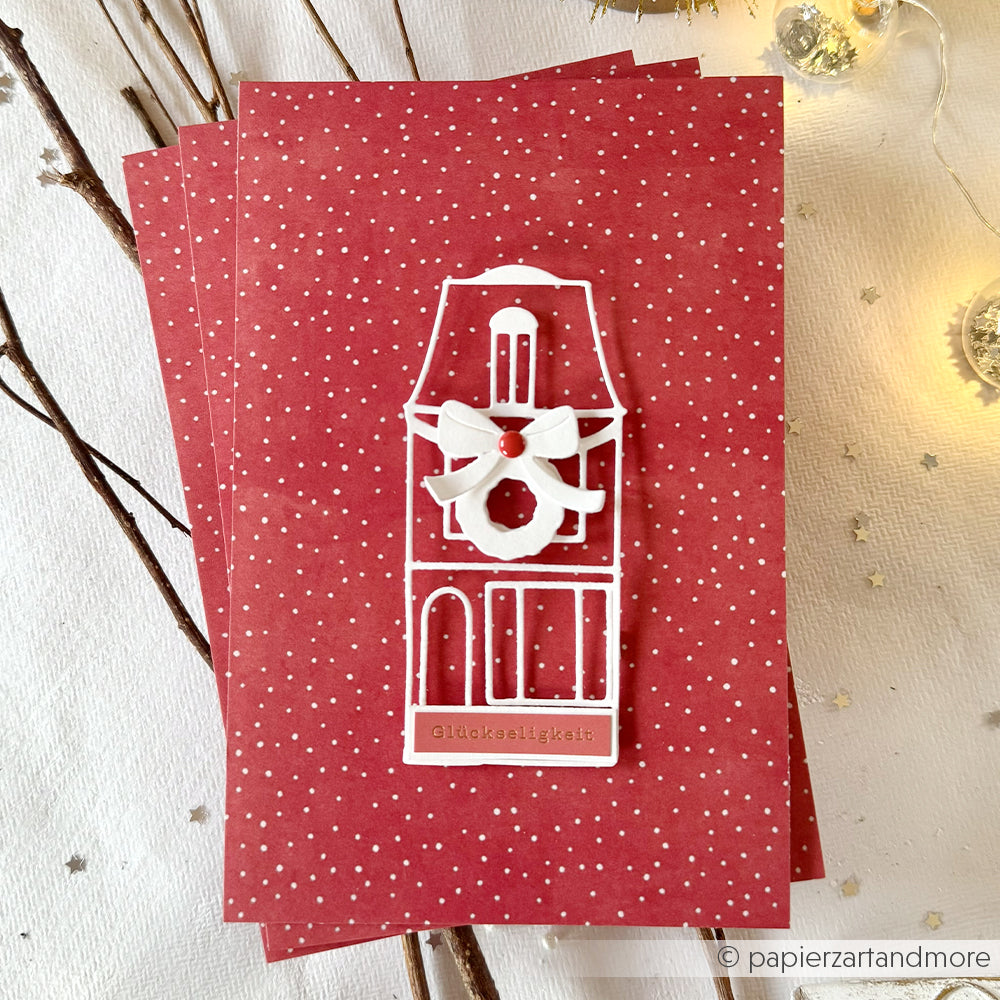 Rote Karte von Alexandra Renke mit weißem Haus und Kranzausschnitt von DieCut Systeme auf einem rot gepunkteten Hintergrund mit Zweigen, Lichterketten und festlichen Designpapieren.