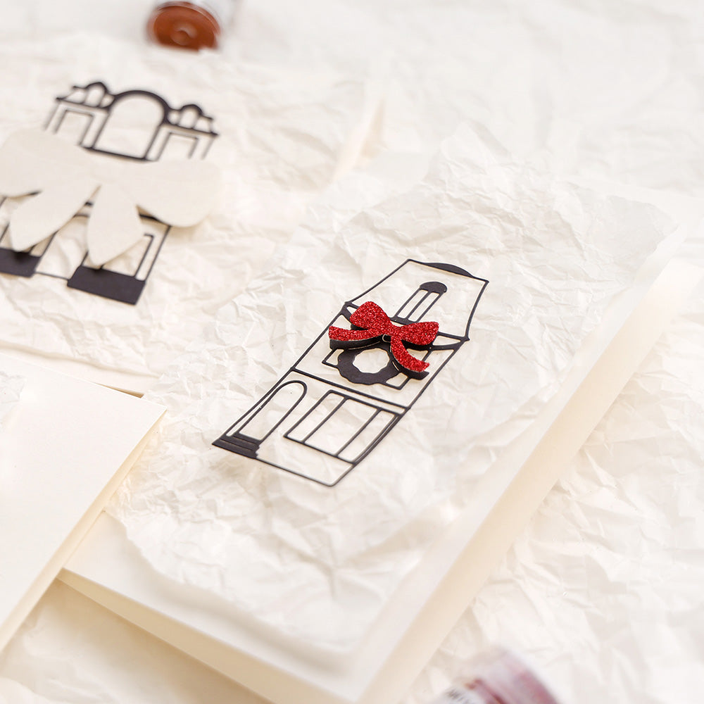 Handgemachte Karte mit schwarzem Gebäudeumriss, Kranz und roter Glitzerschleife auf zerknittertem weißem Designpapier, gestaltet mit der Motivstanze Großes Haus schmal von Alexandra Renke.