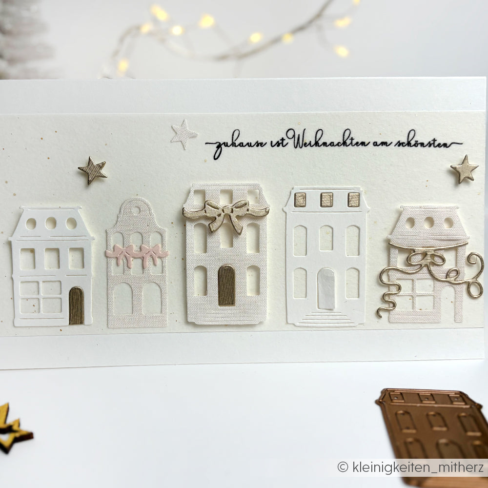 Eine handgefertigte Weihnachtskarte von Alexandra Renke zeigt fünf dekorative Häuser, die mit Designpapier und einer Motivstanze hergestellt wurden, sowie den deutschen Satz "Zuhause ist Weihnachten am schönsten".