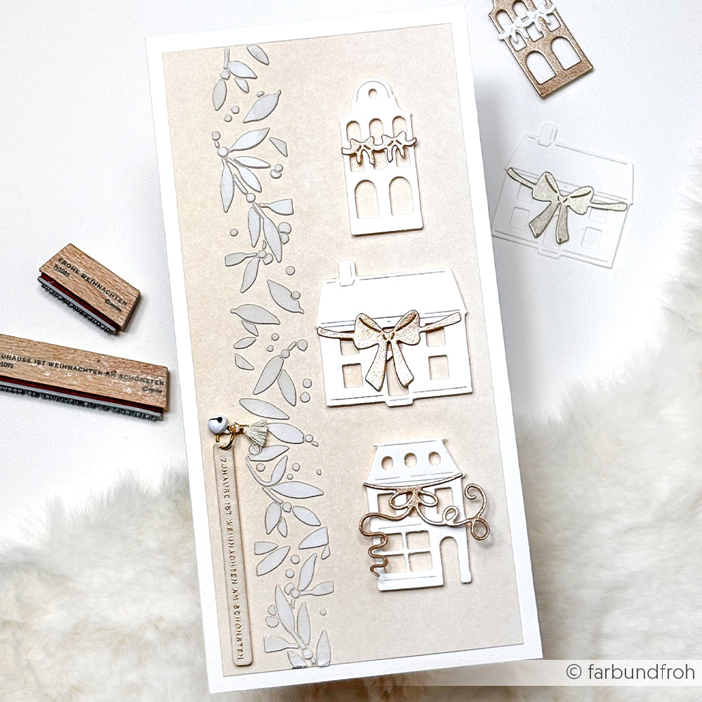 Handgefertigte Karte mit gestanzten Häusern, Blättern und Schleifen von Alexandra Renke auf beigefarbenem Designpapier, zusammen mit Stempeln auf einer weißen, flauschigen Oberfläche.