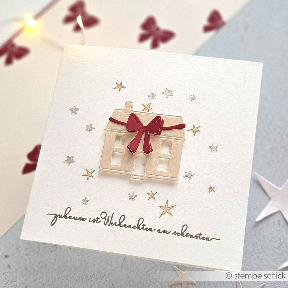 Weiße Karte mit einem Papierhaus mit roter Schleife, Sternenakzenten und deutschem Text - hergestellt mit Alexandra Renke DieCut Systemen und Motivstanze Gemütliches Schleifenhaus auf hochwertigem Designpapier für einzigartige Effekte.