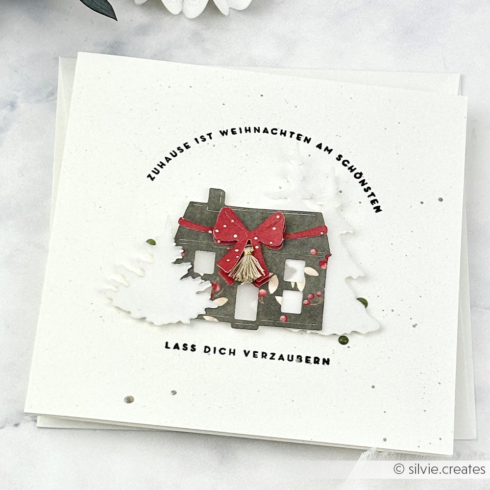 Handgefertigte Weihnachtskarte von Alexandra Renke mit Haus, roter Schleife und deutschem Text ober- und unterhalb des Hauses, hergestellt mit feinen Designpapieren für einen festlichen Touch.