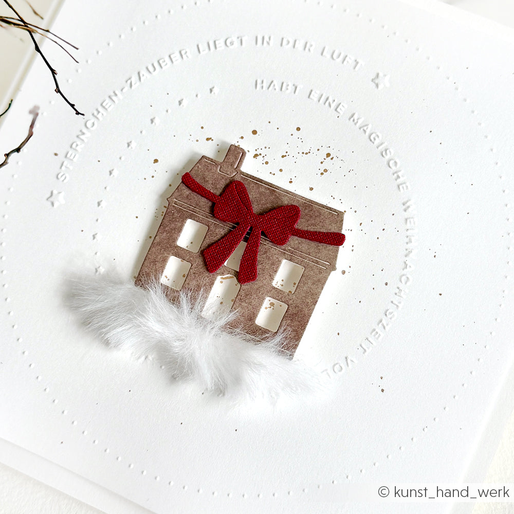Ein Papierhaus mit roter Schleife und weißem Kunstpelz sitzt auf einer weißen Karte, die aus Alexandra Renke Designpapieren gefertigt und mit einem geprägten kreisförmigen Text veredelt ist.