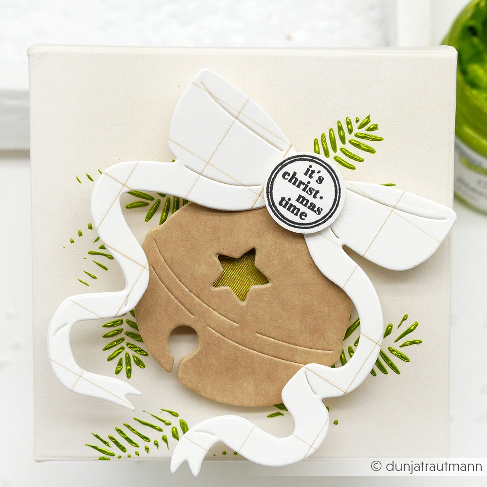 Ein Geschenkkarton mit einer weißen Schleife, einer braunen Glockenverzierung aus der Alexandra Renke Motivstanze Fröhliche Sternenglocke und grünen Tannenzweigen.