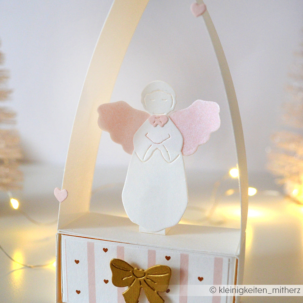Handgemachte Engelsdekoration aus weißem und rosafarbenem Papier von Alexandra Renke, hergestellt mit Designerpapier und Stanzen, mit einer goldenen Schleife, dezenten Lichtern und weichen Bäumen im Hintergrund.