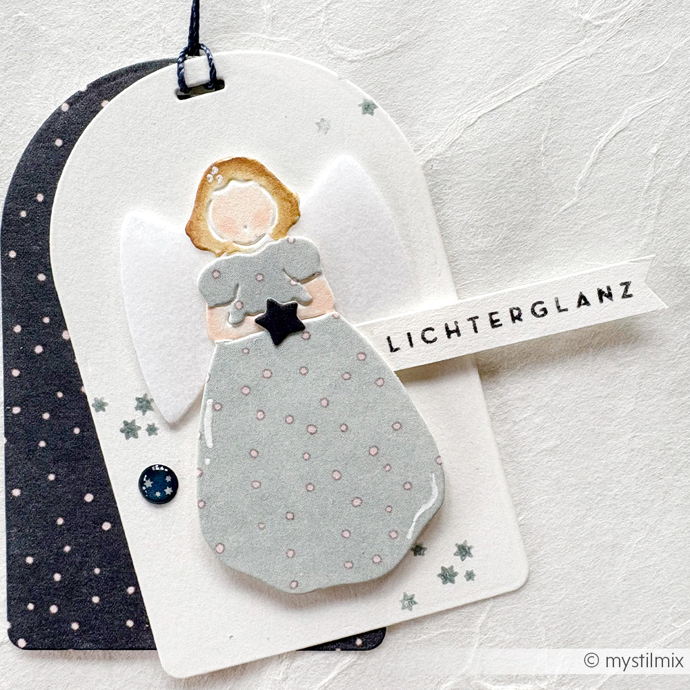 Engel aus Papier von Alexandra Renke, mit grauem Kleid, weißen Flügeln und einem "LICHTERGLANZ"-Anhänger. Hergestellt mit feinen Designpapieren und detailliert mit der Motivstanze Singender Engel.