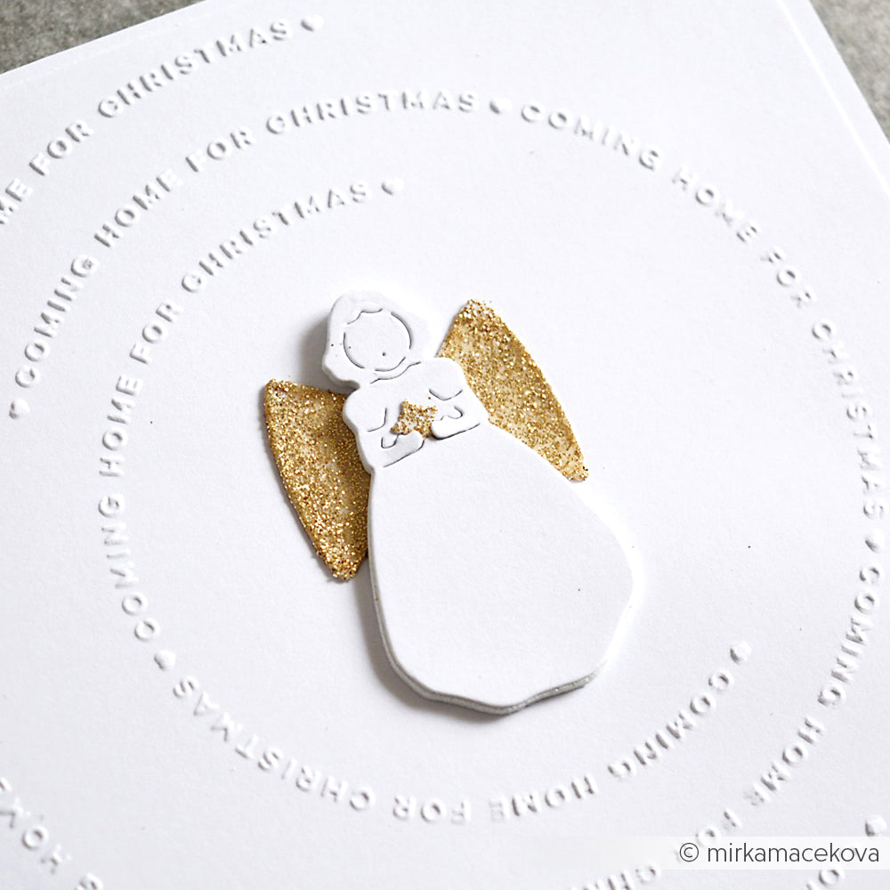 Weißer Papierengel mit der Alexandra Renke Singender Engel Stanze und goldenen Glitzerflügeln auf weißem Karton, mit kreisförmiger Prägung "Coming home for Christmas". Ideal für festliche Designpapiere und Stanzsysteme Projekte.