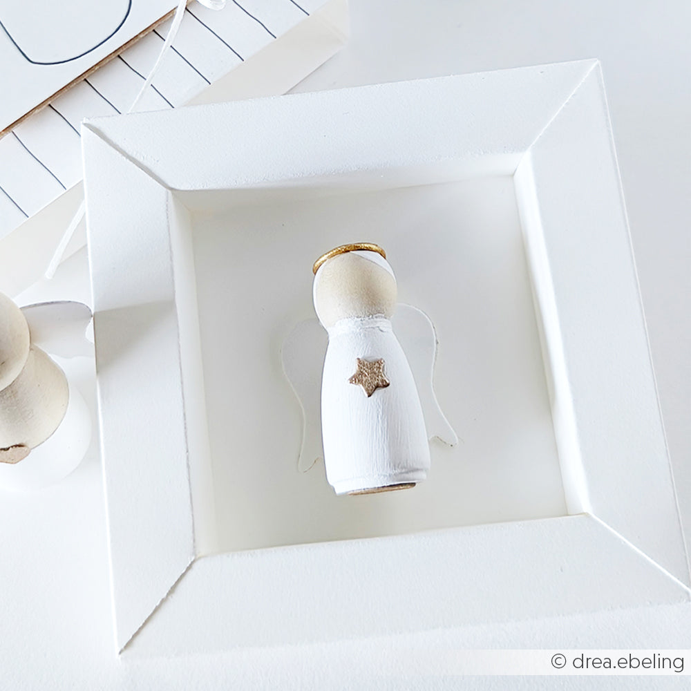 Eine kleine hölzerne Engelsfigur in einem weißen Schattenboxrahmen auf einer weißen Oberfläche, akzentuiert mit zartem Alexandra Renke Designpapier für zusätzlichen Charme.