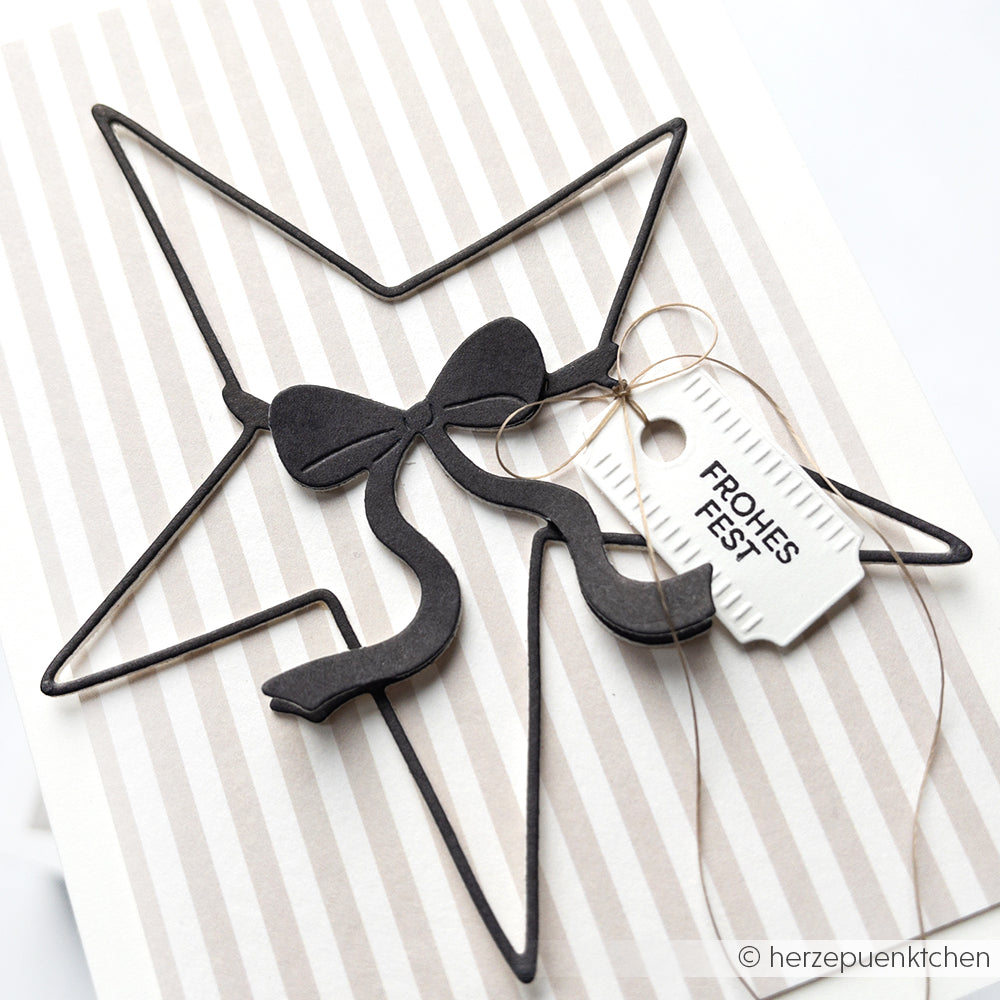 Ein schwarzer Stern und eine Schleife, die mit der Stanze Großer Stern von Alexandra Renke gestaltet wurden, mit einem weißen "Frohes Fest"-Anhänger auf gestreiftem Hintergrund.