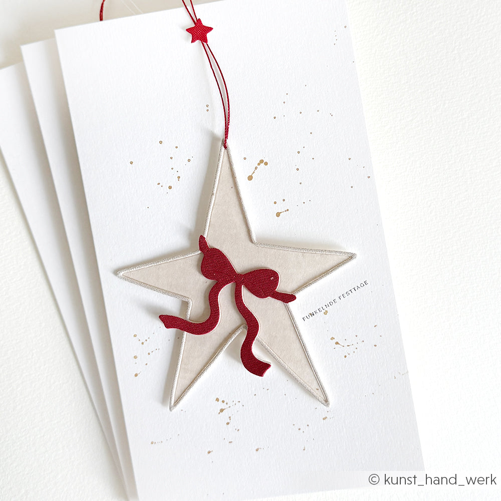 Eine weiße Karte von Alexandra Renke aus eleganten Designpapieren zeigt ein mit der Motivstanze Großer Stern ausgeschnittenes Stern-Ornament und eine rote Schleife, die mit roter Schnur gebunden ist, auf schlichtem weißem Hintergrund.