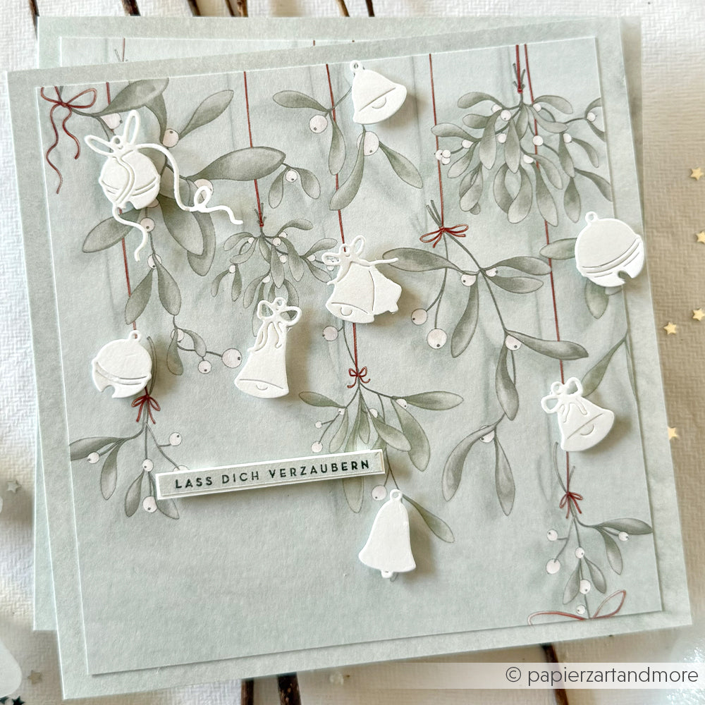 Handgefertigte Karte von Alexandra Renke mit Designpapier, Mistelzweigen, weißen Ornamenten und dem Text "LASS DICH VERZAUBERN" auf hellem Hintergrund, veredelt mit Motivstanze Glöckchen für festlichen Charme.