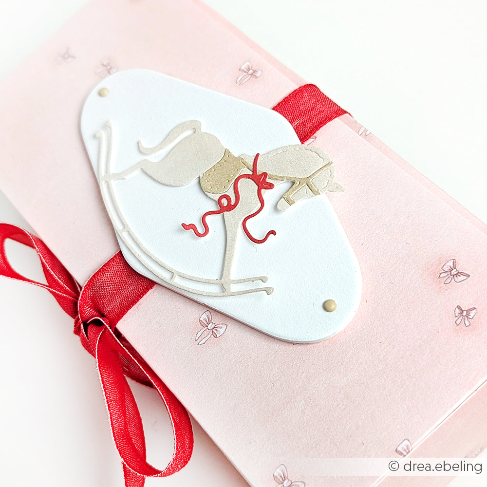 Rosa Umschlag mit roter Schleife und weißem Anhänger mit Schaukelpferd-Illustration, hergestellt mit der Alexandra Renke Motivstanze Schaukelpferd. Ideal für Geschenke oder besondere Anlässe, besonders in Kombination mit Ihren Lieblingsdesignpapieren.