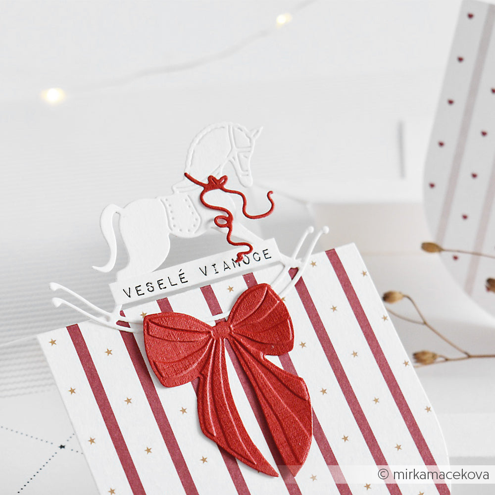 Weiße Karte mit roten Streifen, einer großen roten Schleife und einem weißen Schaukelpferd, ausgestanzt mit der Alexandra Renke Motivstanze Schaukelpferd. Mit dem Text Veselé Vianoce - perfekt für festliche Projekte mit Stanzsystemen.