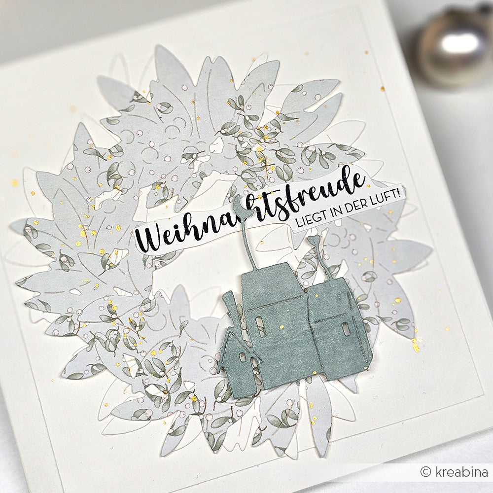 Ein Papierkranz und Häuser, hergestellt mit Alexandra Renke Designpapiere und Motivstanze Mondhäuser, zeigen "Weihnachtsfreude liegt in der Luft!" auf einer weißen Karte.
