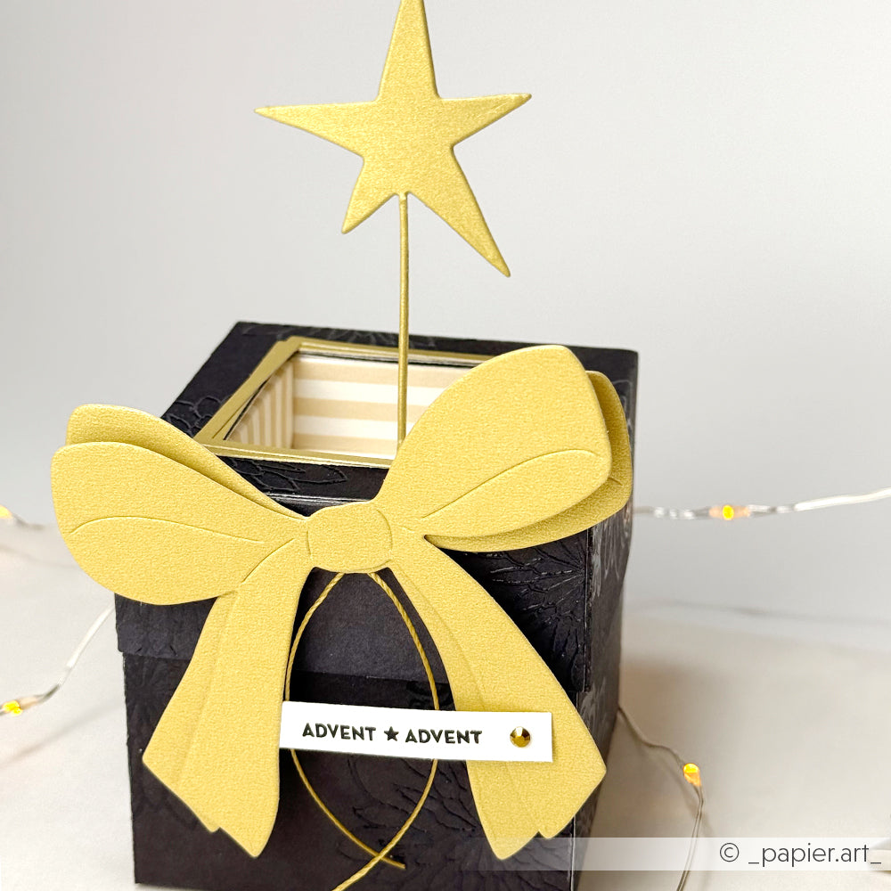 Ein schwarzer Geschenkkarton mit einer großen goldenen Schleife und einem Stern mit der Aufschrift ADVENT ★ ADVENT auf der Vorderseite, mit Alexandra Renke Designpapieren und einer atemberaubenden Motivstanze Sterne für eine festliche Note.