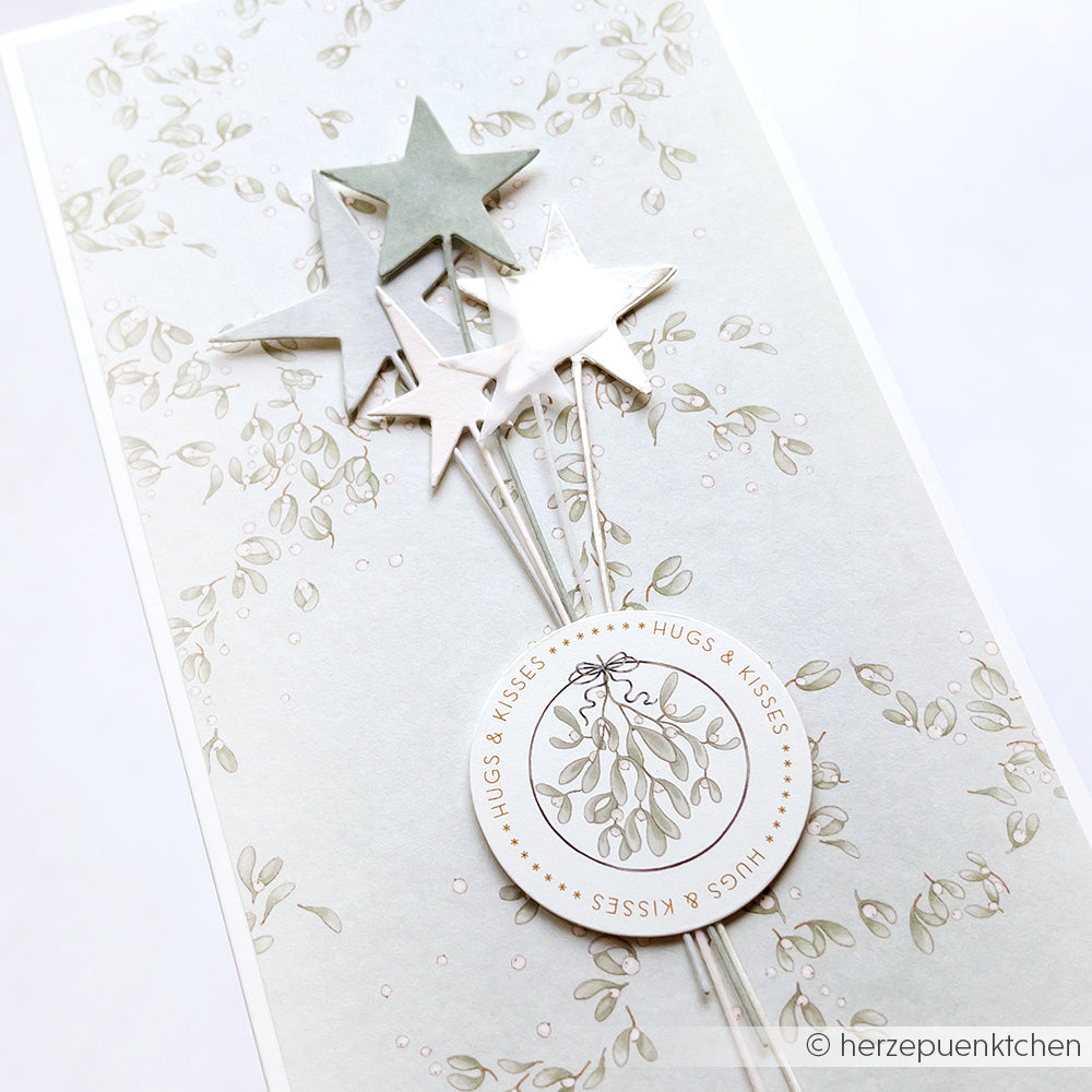 Grußkarte von Alexandra Renke mit einem silbernen Sterne am Stiel-Trio, einem kreisförmigen Hugs & Kisses-Etikett und einem eleganten Blatthintergrund aus hochwertigem Designpapier.