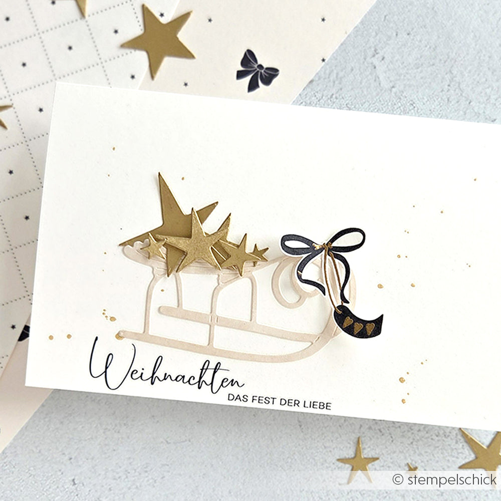 Weiße Karte von Alexandra Renke mit einem goldenen Schlitten, Sternstanzung und schwarzer Schleife auf hochwertigem Designpapier. Text: Weihnachten DAS FEST DER LIEBE.