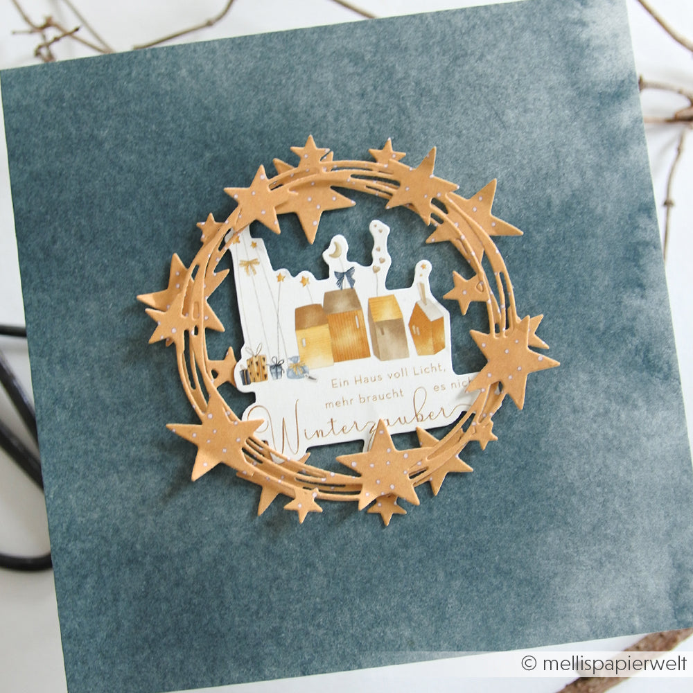 Handgefertigte Weihnachtskarte von Alexandra Renke mit Designpapieren und einem mit der Motivstanze Fröhlicher Sternenkranz ausgeschnittenen Sternenkranz, sowie einer gemütlichen Dorfszene auf strukturiertem blauem Hintergrund.
