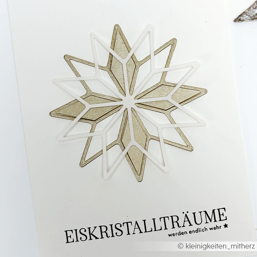 Eine geometrische Schneeflocke in Gold und Weiß, gestaltet mit Alexandra Renke Designpapieren und der Motivstanze Achterstern, über dem deutschen Text "Eiskristallträume werden endlich wahr.