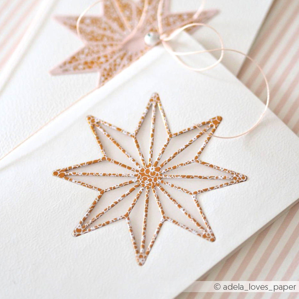 Weiße Karte von Alexandra Renke mit einem verschlungenen goldenen Stern, der mit der Motivstanze Achterstern ausgestanzt wurde, auf einem rosa und weiß gestreiften Hintergrund.