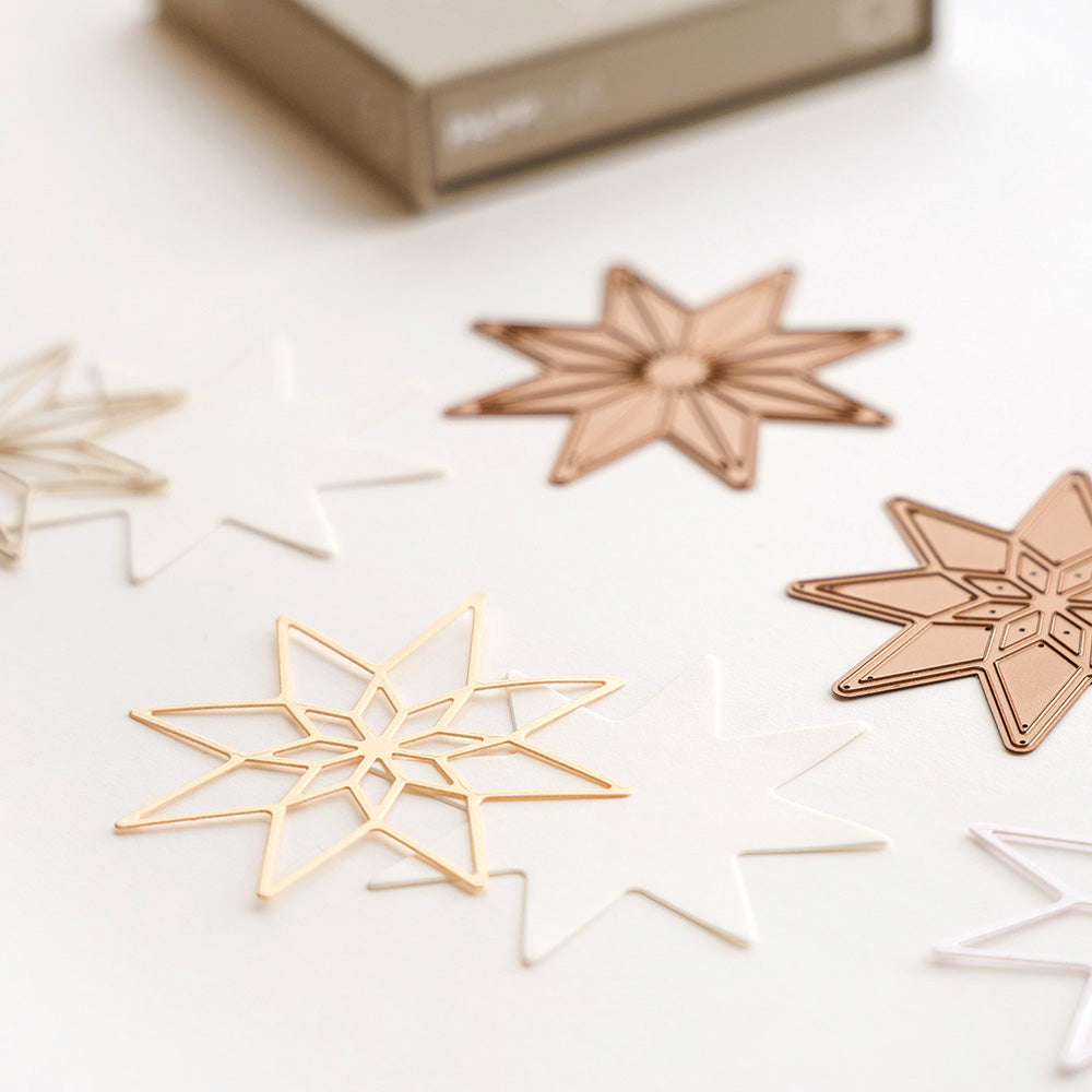 Gold-, weiß- und kupferfarbene sternförmige Papierausschnitte, die mit Alexandra Renke Designpapieren und der Motivstanze Achterstern Strahlen Innenstern hergestellt wurden, sind auf einer hellen Fläche angeordnet, mit einer Box im Hintergrund.