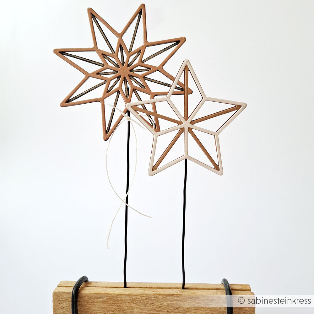 Zwei Metallstern-Ornamente von Alexandra Renke, gefertigt mit Motivstanze Achterstern und auf schwarzen Drahtstielen, stehen auf einem Holzblock vor weißem Hintergrund.