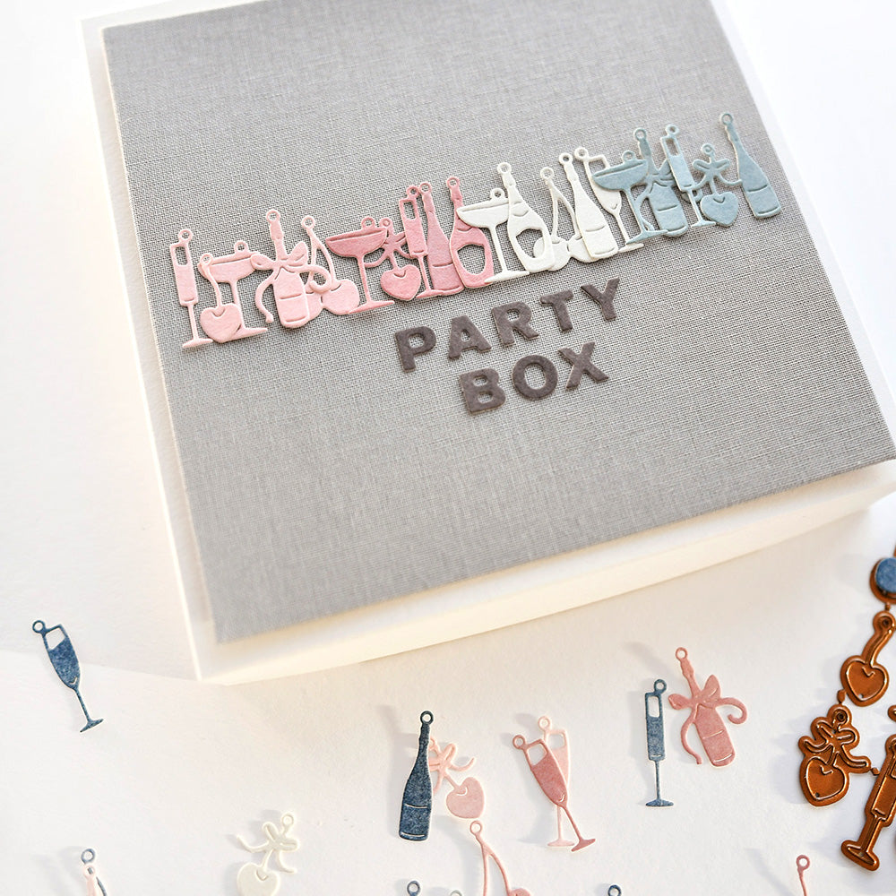 Eine graue Alexandra Renke Party Box mit bunten Designpapieren und Fröhliche Drinks Anhängern auf dem Deckel und rundherum verstreut.