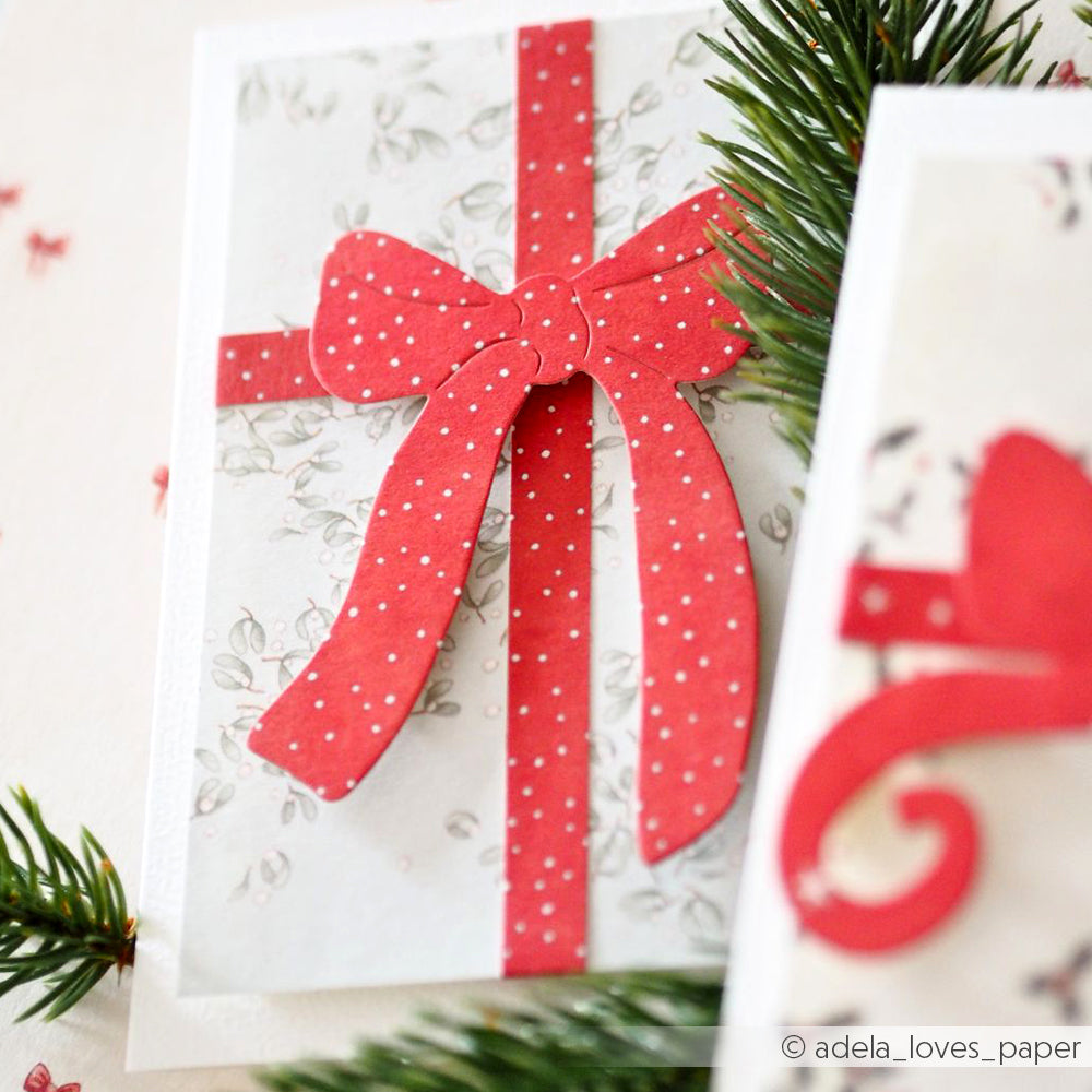 Handgemachte Karte von Alexandra Renke mit einer roten Tupfenschleife aus der Motivstanze Edelschleife auf weißem Hintergrund mit Tannenzweigen in der Ecke.