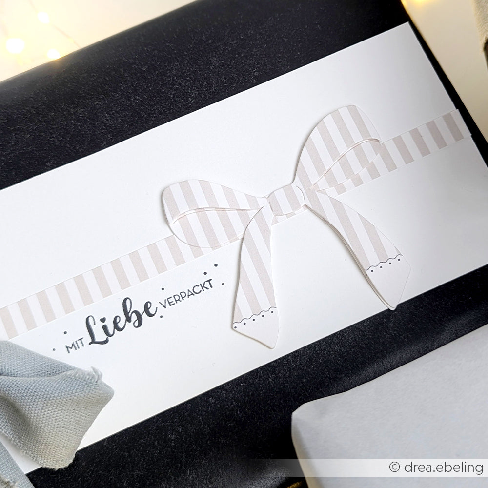 Weißer Geschenkanhänger mit gestreifter Papierschleife, hergestellt mit der Motivstanze Präsentschleife von Alexandra Renke, mit dem deutschen Text "Mit Liebe verpackt" auf einem schwarz verpackten Geschenk.