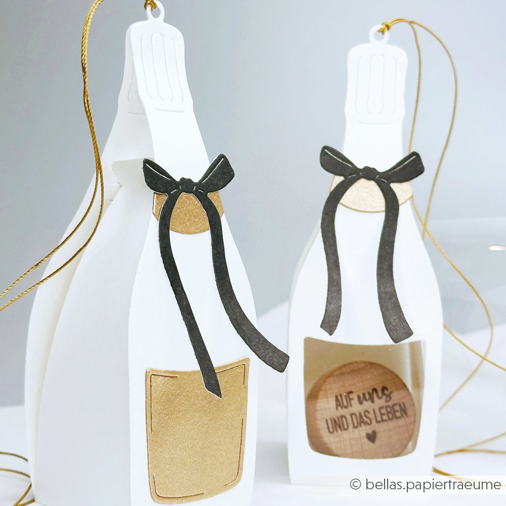 Zwei weiße Sektflaschen-Ornamente aus Papier von Alexandra Renke sind mit goldenen Etiketten und schwarzen Schleifen versehen und aus Designpapieren gefertigt. Ein Ornament enthält eine spezielle deutsche Nachricht im Inneren für ein besonders liebevolles Detail.