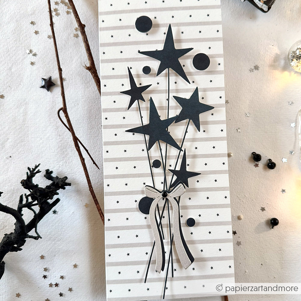 Handgemachte Karte von Alexandra Renke mit schwarzen Sternen an Schnüren und einer Schleife, gefertigt mit der Motivstanze Feinschleife, auf Designpapieren mit gepunktetem, gestreiftem Hintergrund.