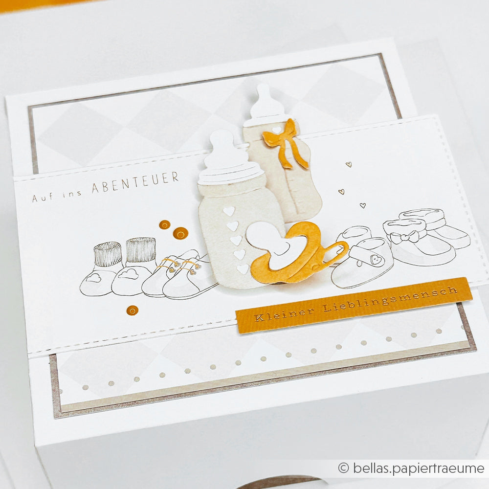 Eine Karte zum Thema Baby von Alexandra Renke mit Fläschchen, Schnuller und Schuhen auf weiß-goldenem Hintergrund, gestaltet mit Designpapieren und dem Motivstanze Fläschchen Set.