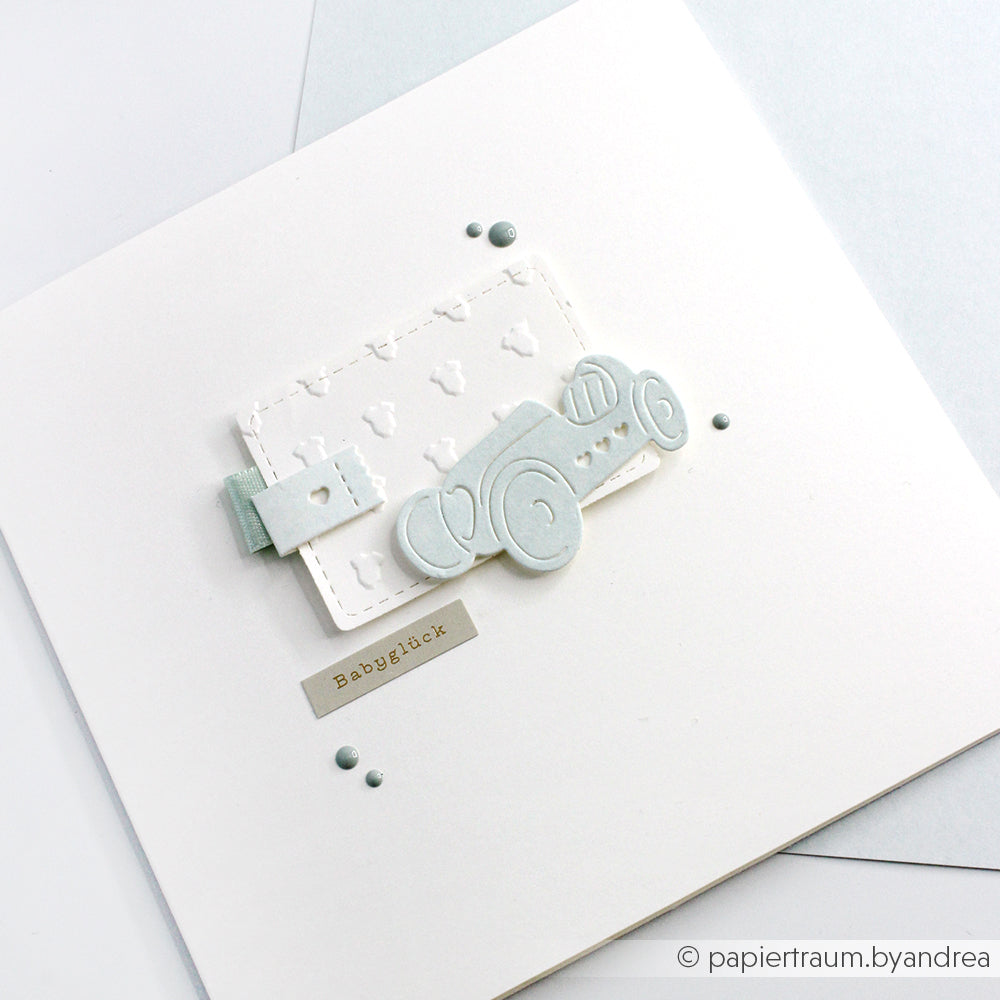 Weiße Karte mit hellblauem Babyauto, strukturiertem Anhänger aus Designpapieren und "Babyglück" auf neutralem Hintergrund - gestaltet mit der Motivstanze Seifenkisten Trio von Alexandra Renke und Stanzsysteme.
