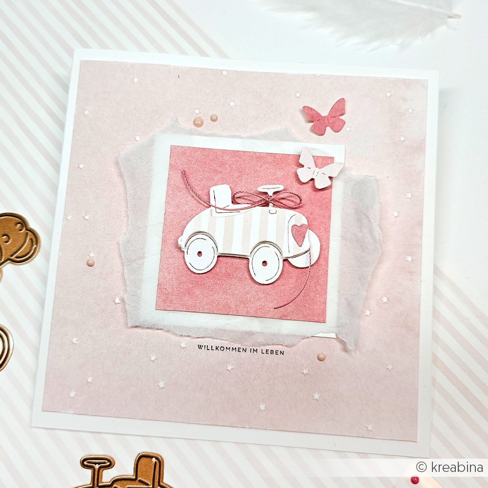 Eine handgefertigte Karte von Alexandra Renke mit einem Spielzeugauto und Schmetterlingen mit rosa Akzenten und dem Text "Willkommen im Leben" darunter. Hergestellt mit Motivstanze Seifenkisten Trio und hochwertigen Designpapieren für einen charmanten, detaillierten Look.