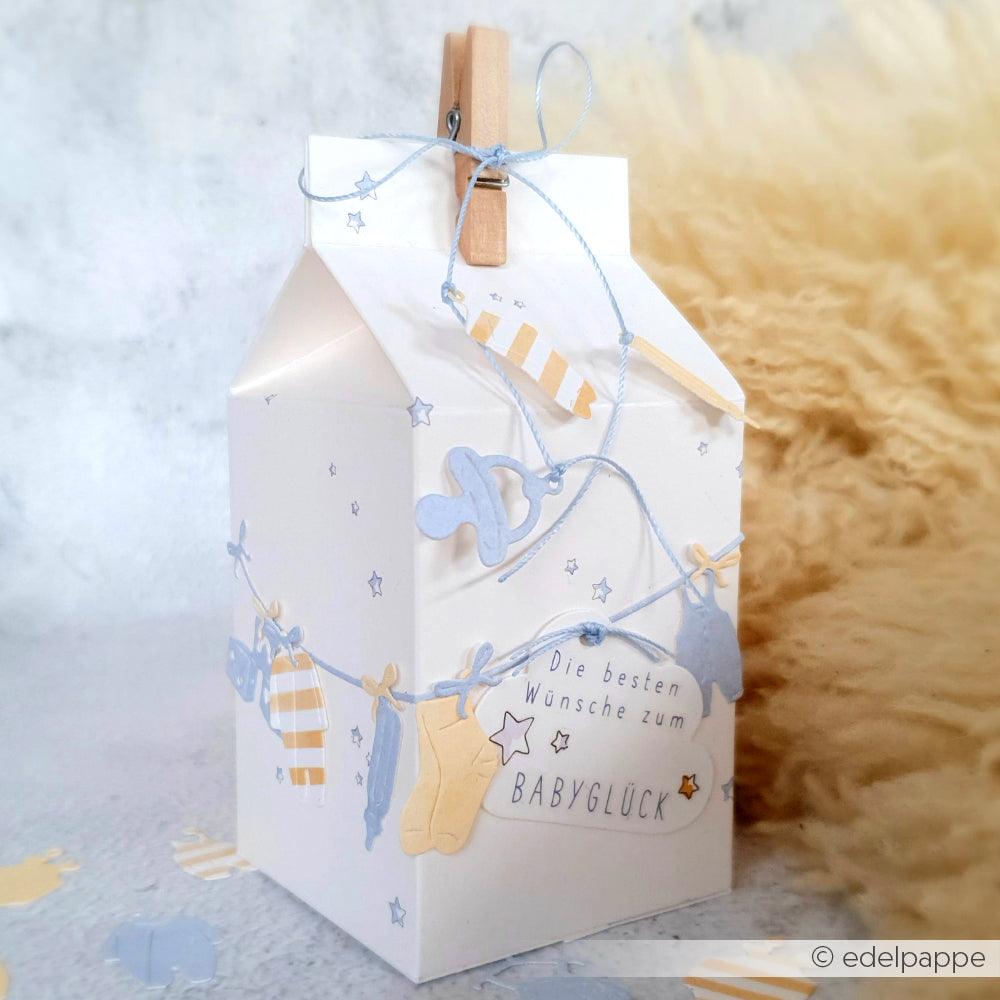 Geschenkbox von Alexandra Renke, dekoriert mit Babykleidung, Schnullern, einem Zettel "Die besten Wünsche zum BABYGLÜCK", Babyanhänger-Ausstanzungen und eleganten Designerpapieren für einen liebevollen Look.