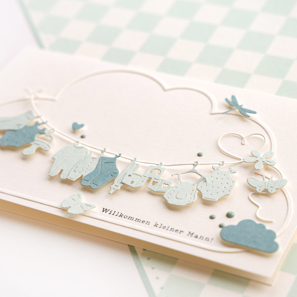 Die Alexandra Renke Babykarte zeigt eine Wäscheleine mit kleinen Kleidungsstücken, Schmetterlingen und dem Spruch "Willkommen kleiner Mann!", gefertigt aus hochwertigen Designpapieren und veredelt mit einer Motivstanze Baby Anhänger.