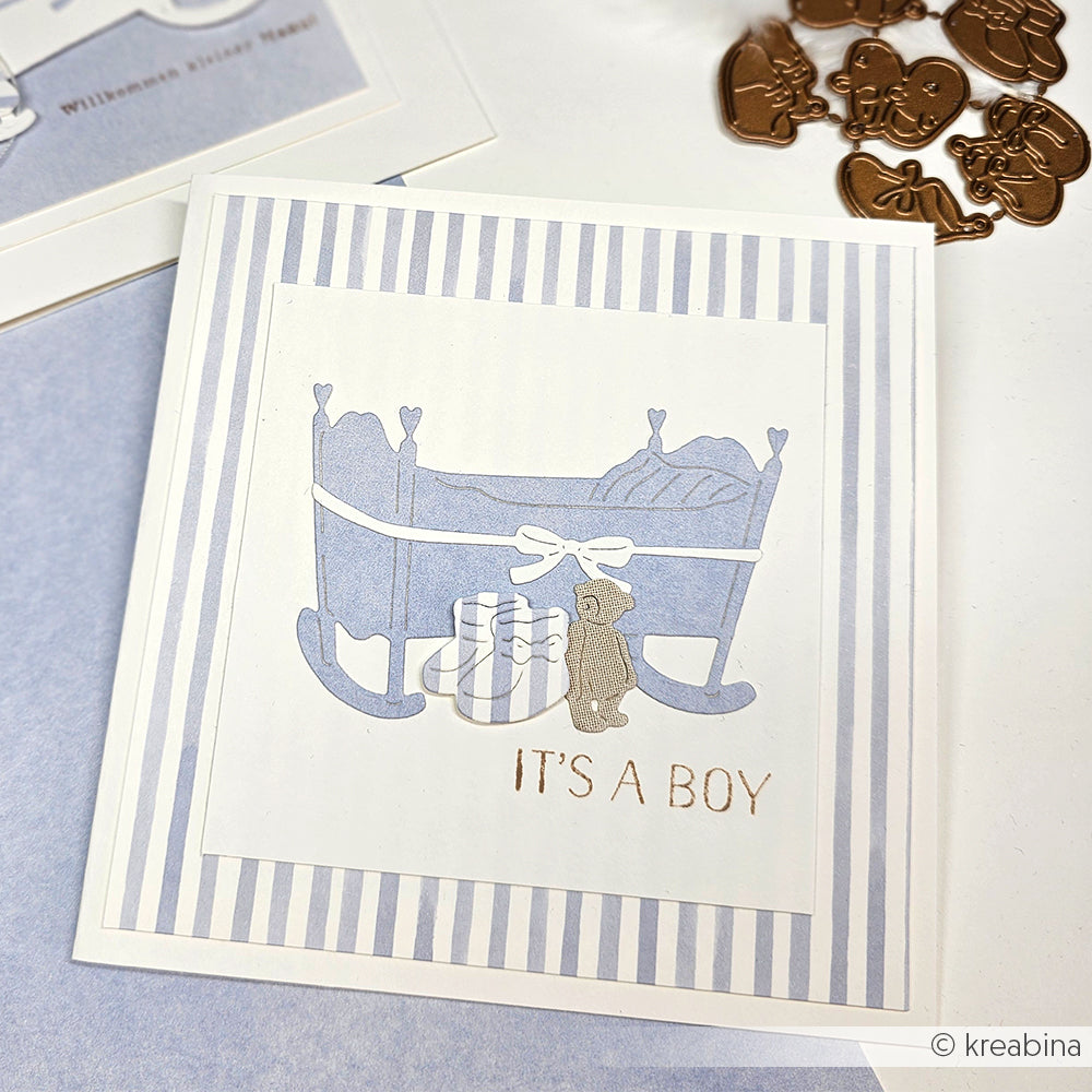 Grußkarte von Alexandra Renke mit blauer Wiege, Geschenken, Teddybär und "ITS A BOY" auf gestreiftem Hintergrund - gestaltet mit Motivstanze Herzliche Wiege und hochwertigen Designpapieren.