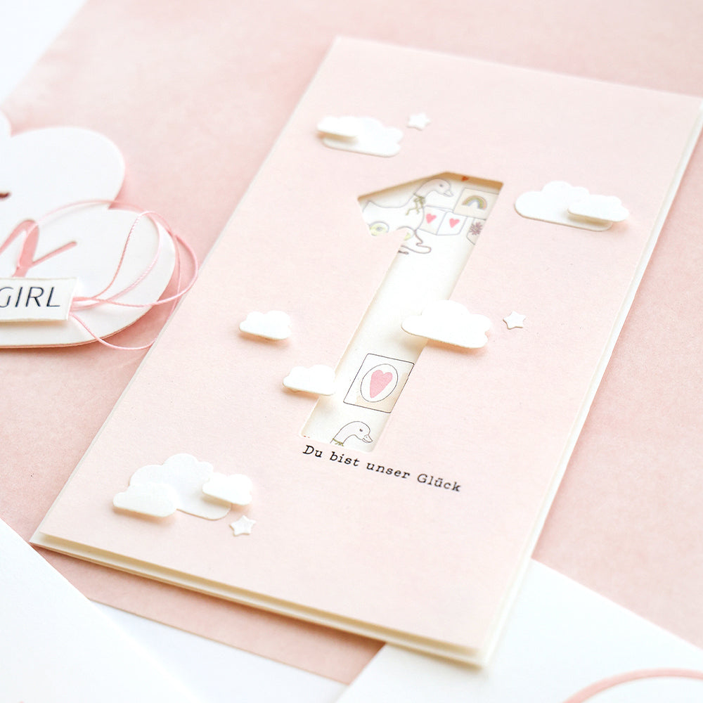 Rosafarbene Karte von Alexandra Renke mit Wolken, einer mit Motivstanze ausgeschnittenen Nummer 1 und dem Text "Du bist unser Glück" auf rosa Hintergrund.