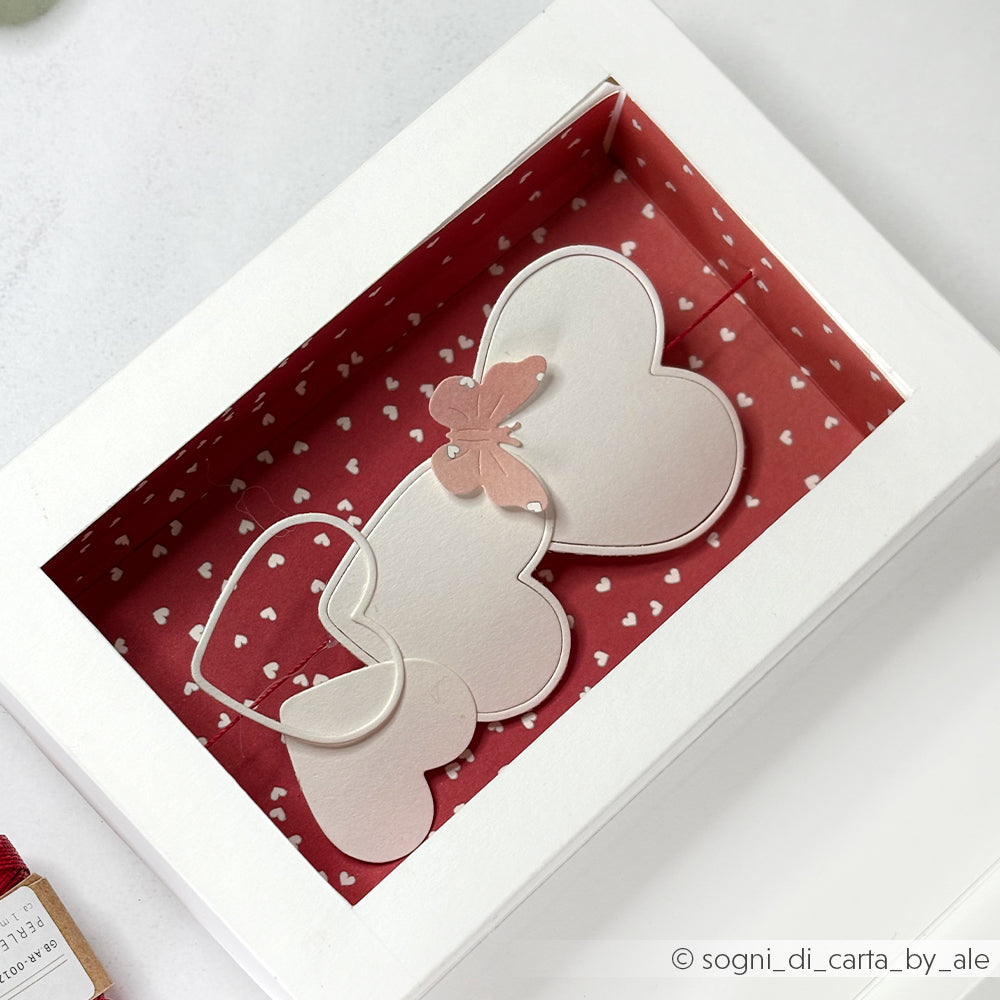 Eine weiße Schachtel mit herzförmigen Karten aus Designpapieren von Alexandra Renke, verziert mit einem rosafarbenen Schmetterling, mit einer Motivstanze Herzrahmen, auf rotem Hintergrund mit weißen Herzen.