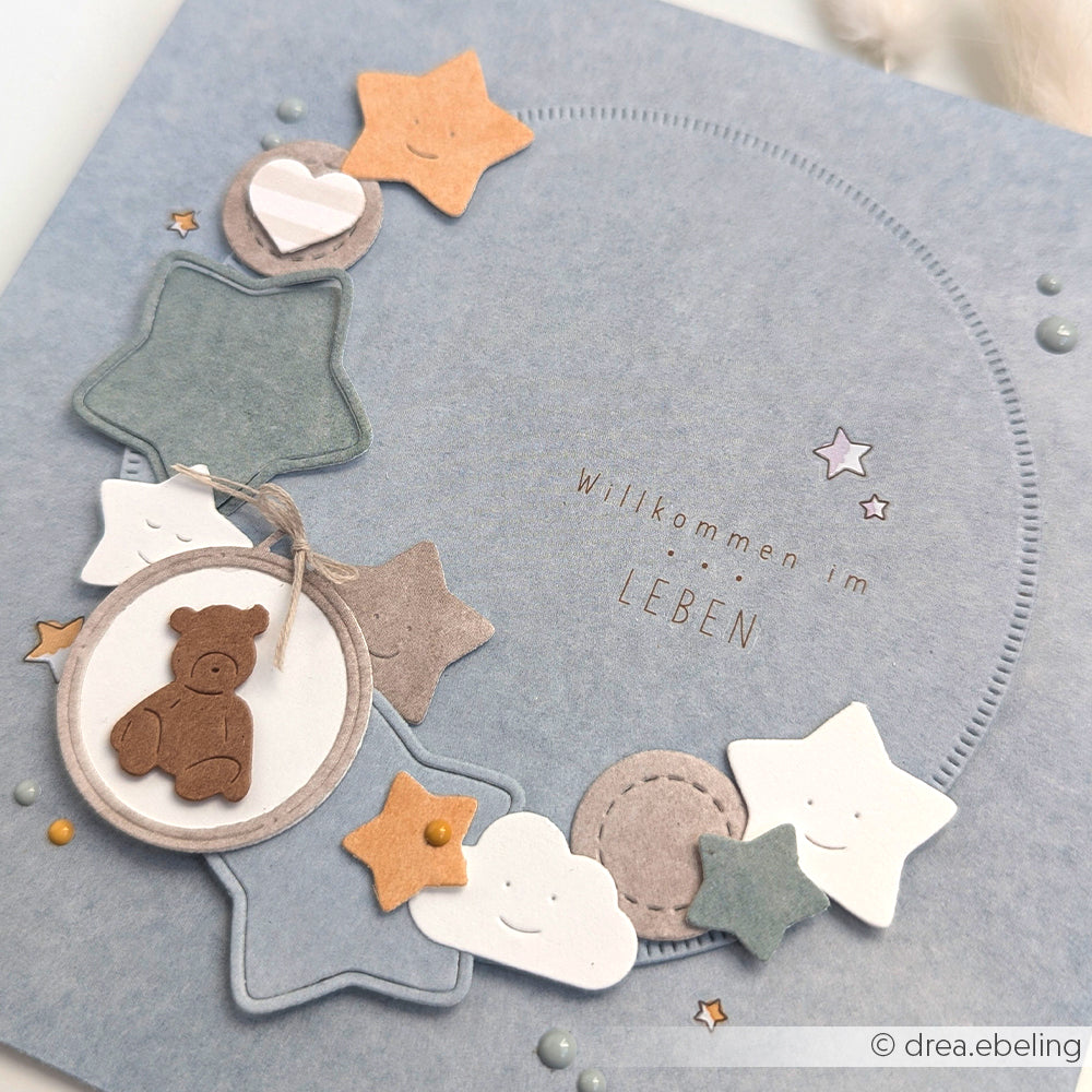Blaue Karte von Alexandra Renke mit Sternen, Wolken und einem Teddybär-Charme aus Designpapier; Text: "Willkommen im Leben.